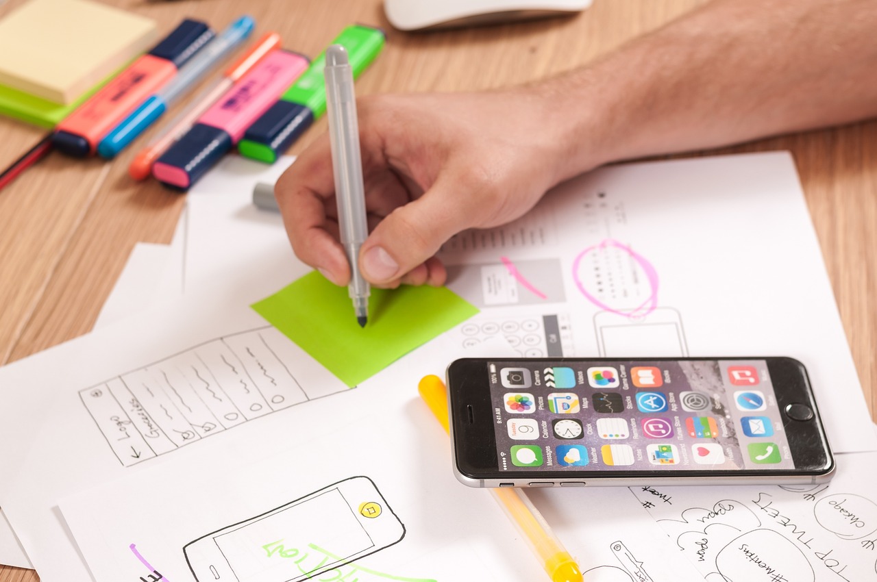 Person sketching mobile UI layouts surrounded by planning notes and a smartphone, representing strategic design and speed optimization for Shopify to improve user experience during peak shopping seasons.