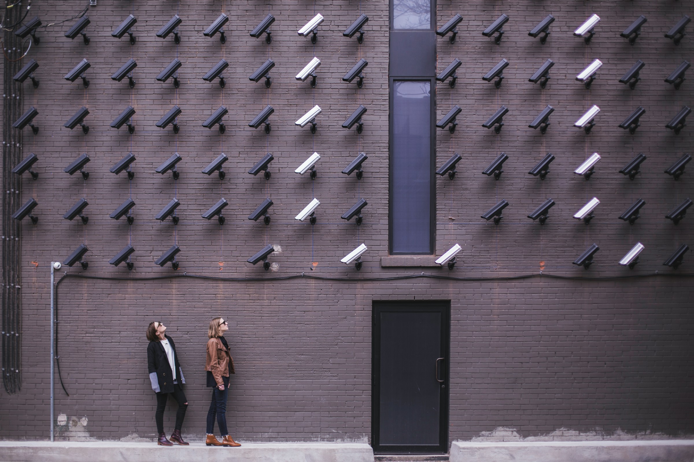 Tracking Pixels That Convert: How to Use Them Effectively 13 Two women standing under a wall covered with dozens of surveillance cameras, symbolizing online tracking, spy pixels, and data privacy concerns.