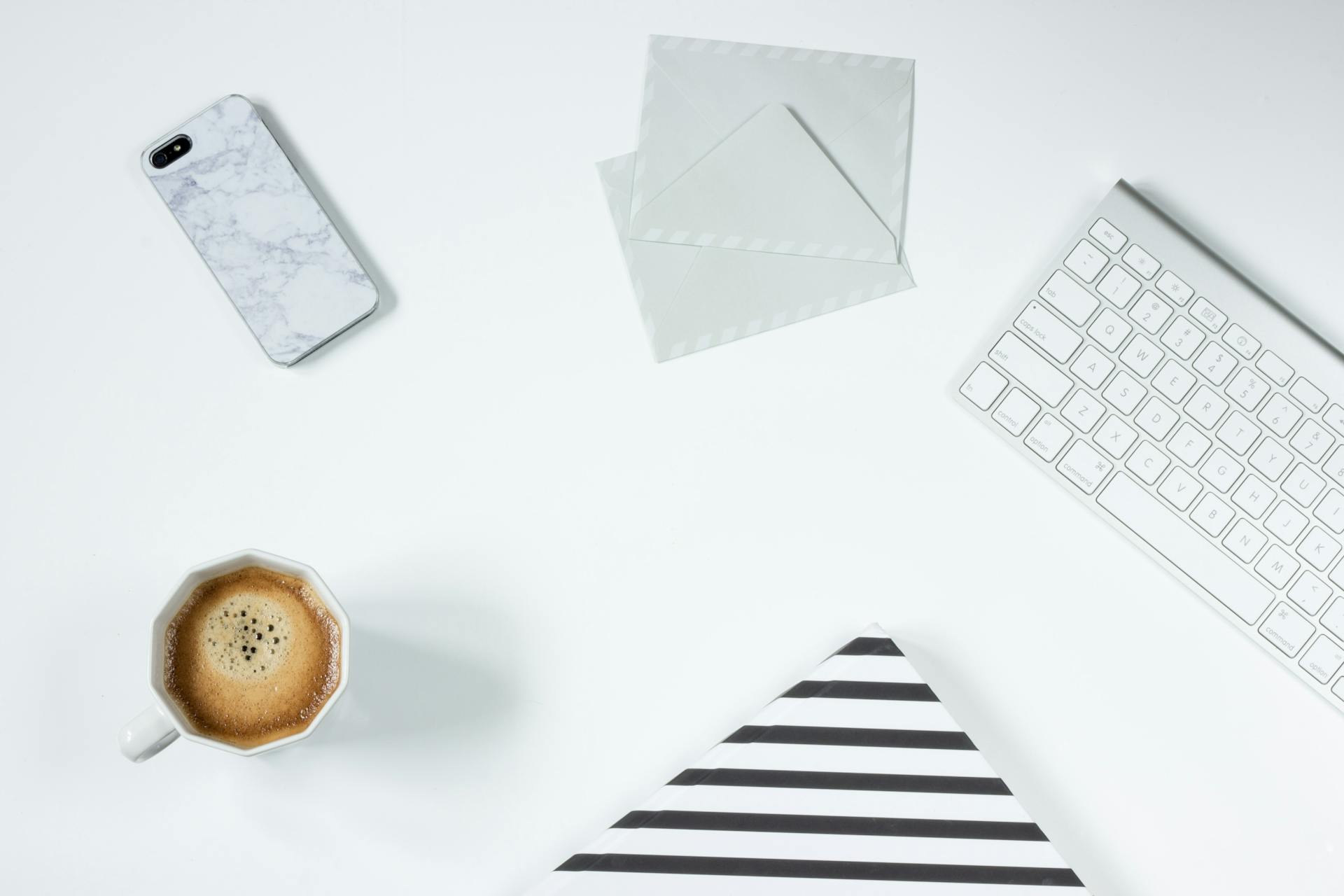 Flat lay of a modern white desk with coffee, smartphone, envelopes, and keyboard, representing digital and physical communication in ecommerce email marketing workflows.