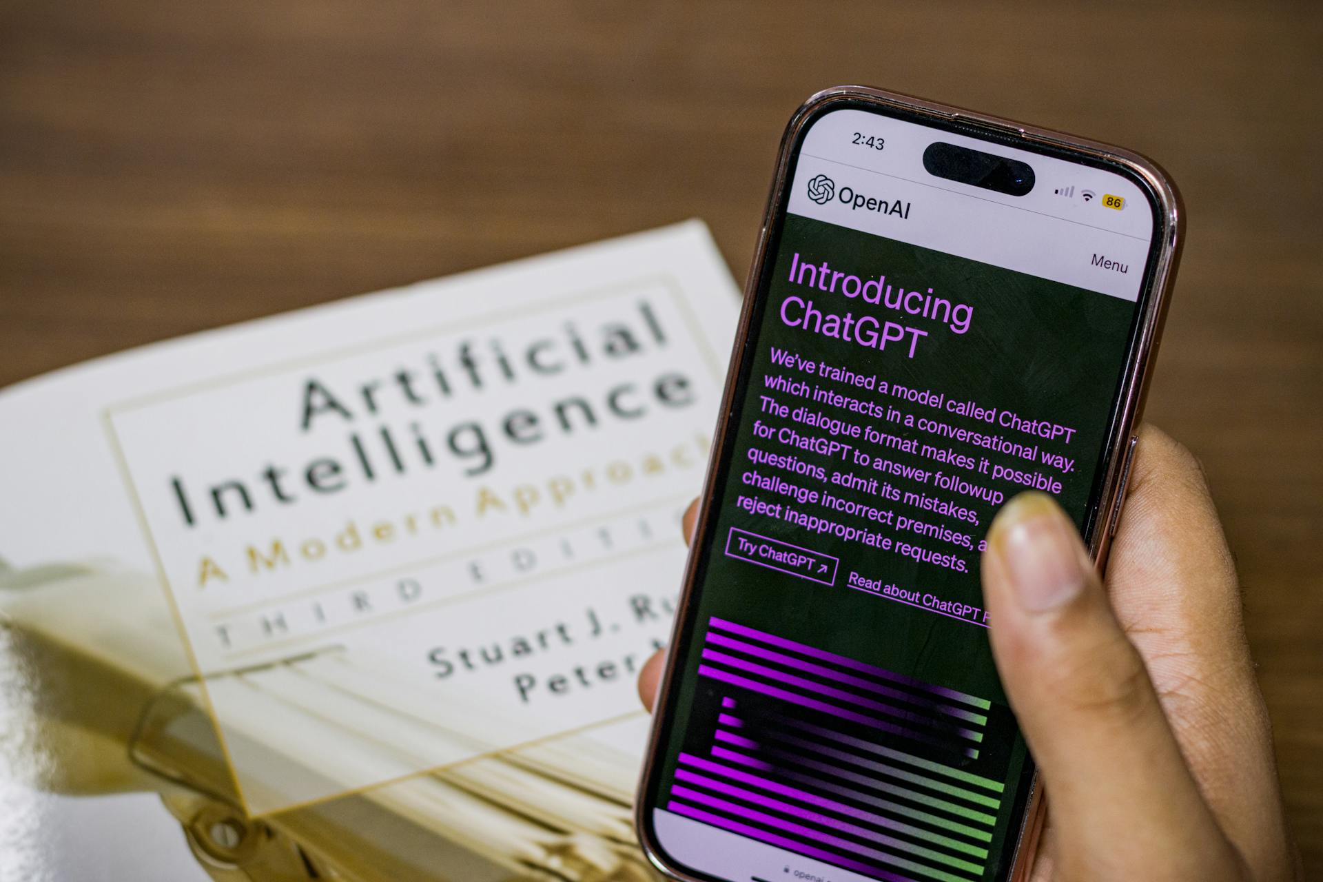 Person holding a smartphone displaying the ChatGPT introduction screen, with an AI textbook in the background—highlighting the role of generative AI in the AEO vs GEO conversation.