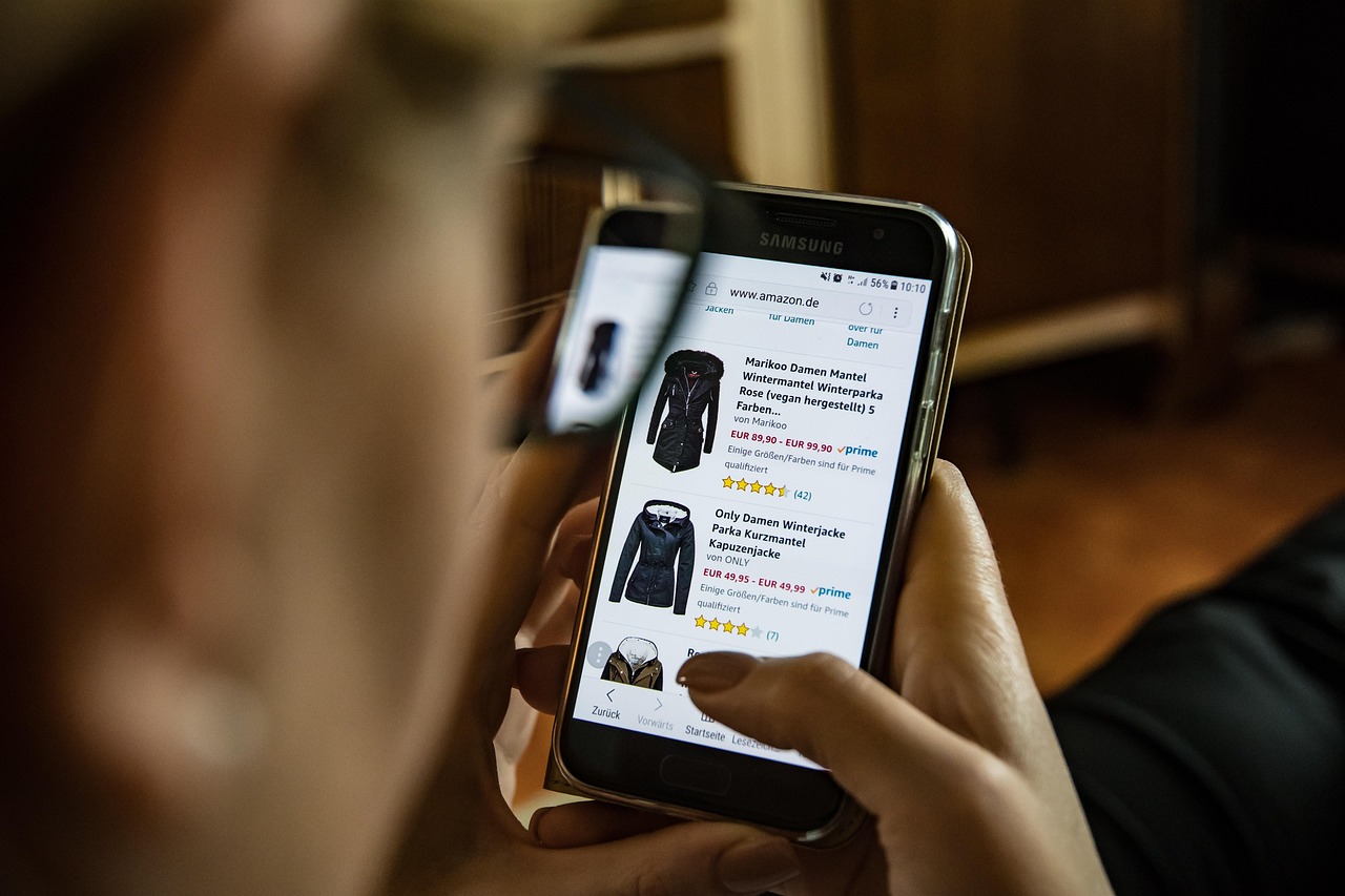 Person browsing winter jackets on Amazon using a smartphone, illustrating mobile-first ecommerce behavior, online shopping trends, and product comparison through reviews and pricing.